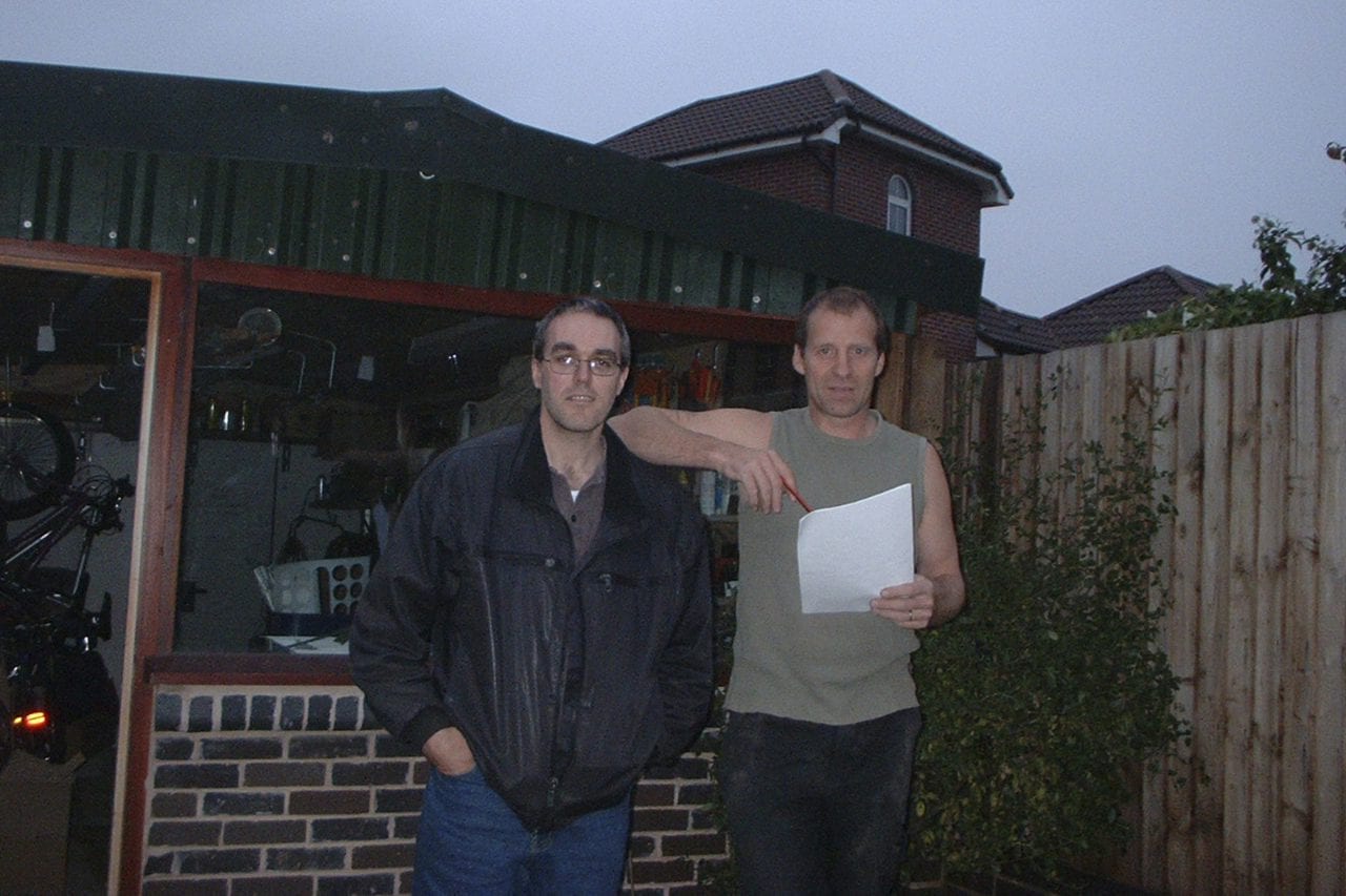 Managing Director Brett Barratt holding a pen and paper standing with another Warrior Employee in front of Bretts old garage, the original Warrior Doors workshop. Photo taken in the 90s.
